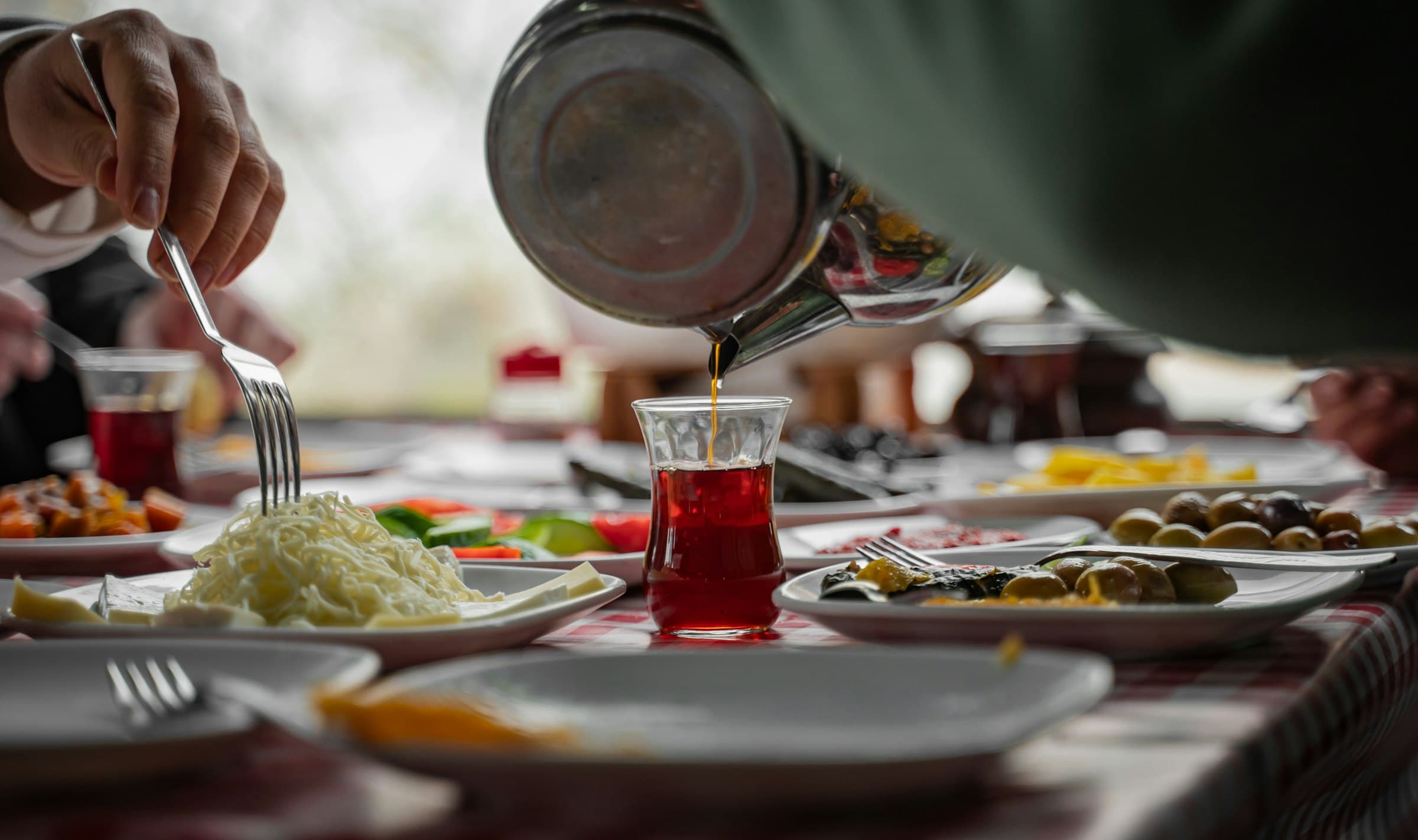 Türkischer Tee wird aus einer Kanne in ein Tulpenglas eingeschenkt, im Hintergrund ein gedeckter Tisch